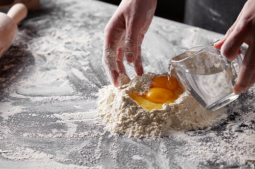 women's hands baker pours water in a pile of flour with yolks on a kitchen table. Step-by-step preparation of the dough