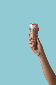 Hand of a girl holding a crispy waffle cone with melting ice cream with colored pills splashes of ice cream flowing on her hand around a blue background with copy space for text