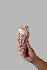 A woman's hand holds melting vanilla ice cream with multi-colored tablets in a waffle cone on a gray background. Copy space for text.