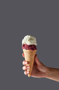 A red fruit ball and a vanilla white ice cream ball in a waffle cone held by a woman's hand against a gray background. Summer concept with copy space.