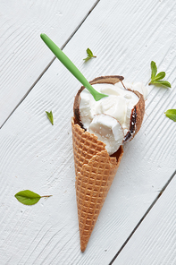 Waffle cone with ice cream in coconut with a spoon and mint leaves On white wooden background flat lay
