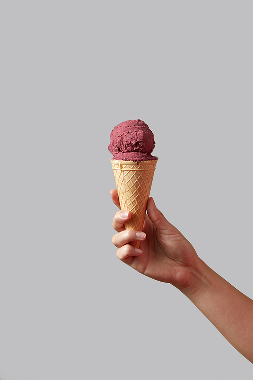 Berry red ice cream in the form of ball in a waffle cone, which is held by a woman's hand on a gray background. Summer concept with copy space.