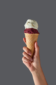 A female hand holds red berry gelato and white milk ice cream in a waffle cone on a gray background. Summer concept with copy space.