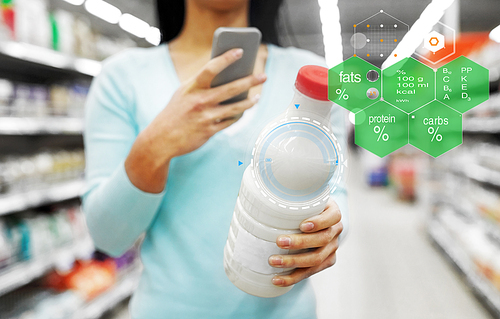 eating, technology and people concept - young woman with smartphone holding milk bottle at grocery store or supermarket over food nutritional value chart