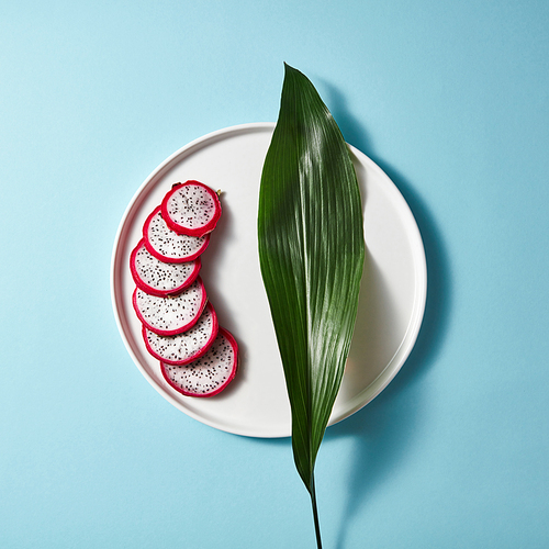 Several pieces of a sliced white pink pitahaya on white plate with green leaf on blue background. Flat lay and copy space