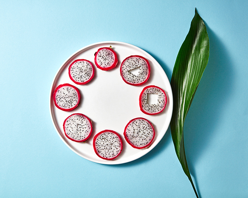 Composition from a white round plate with slices of Pitahaya and a green leaf with reflection of shadows on a blue paper background with copy space. Top view