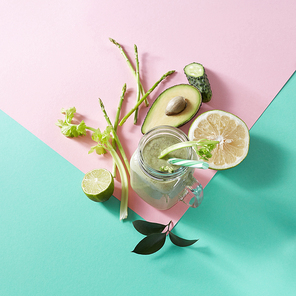 Vegetarian healthy smoothies from green vegetables with green leaves, slices of lemon, avocado, cocumber and plastic straw in a glass bowl on duotone pink green paper background. Top view.
