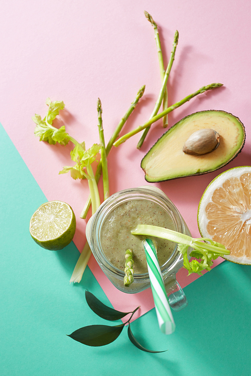 Vegetarian healthy smoothies from green vegetables with green leaves, slices of lemon, avocado, cocumber and plastic straw in a glass bowl on duotone pink green paper background. Top view.