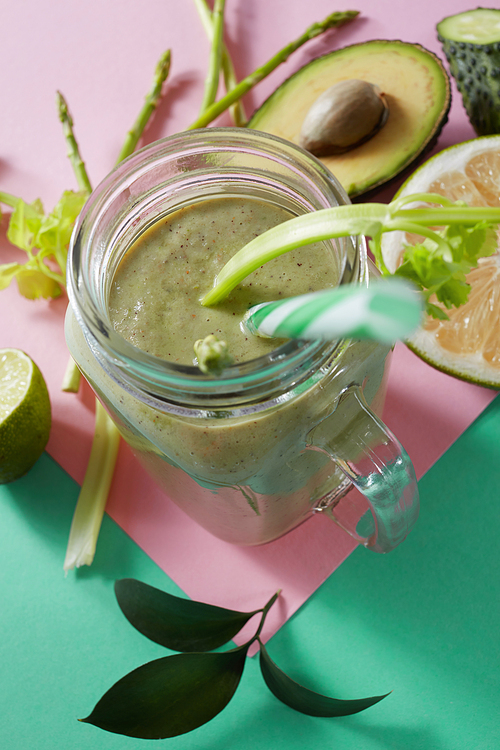 Vegetarian healthy smoothies from green vegetables with green leaves, slices of lemon, avocado, cocumber and plastic straw in a glass bowl on duotone pink green paper background.