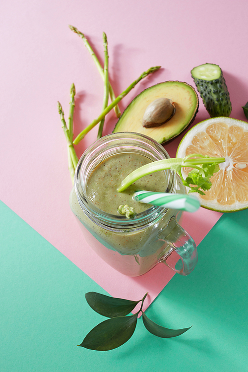 Vegetarian healthy smoothies from green vegetables with green leaves, slices of lemon, avocado, cocumber and plastic straw in a glass bowl on duotone pink green paper background.