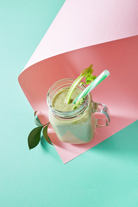 Vegetarian healthy smoothies from green vegetables with green leaves and plastic straw in a glass bowl on duotone pink green paper background. Pink paper twisted in any shape.
