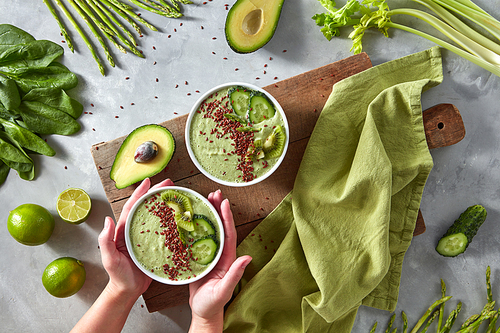 The girl's hands hold a plate of detox smoothies made from green vegetables, kiwi, avocado, cucumber, spinach, lime and flax seeds on a wooden board on a gray kitchen table.