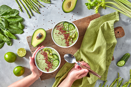 Homemade vegetable smoothies. A woman's hand holds a plate of dietary smoothies from avocados of asparagus with slices of cucumber, kiwi and flax seeds on the background of a wooden board on a gray kitchen table. Healthy food. Top view