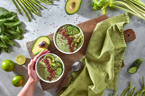 A woman's hand holds a plate of dietary smoothies made from avocado, cucumber, spinach, asparagus, lime with kiwi and flax on a wooden board on a gray kitchen table. Top view