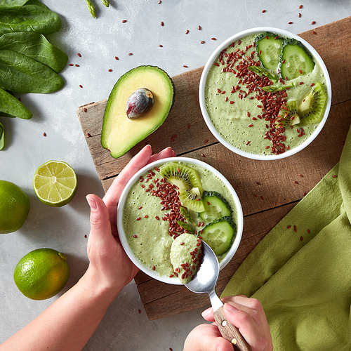 A woman's hand with a spoon takes a diet smoothie. Nutritious smoothies with cucumber, kiwi, avocado, spinach, celery and flax seeds on a gray kitchen counter. Healthy food