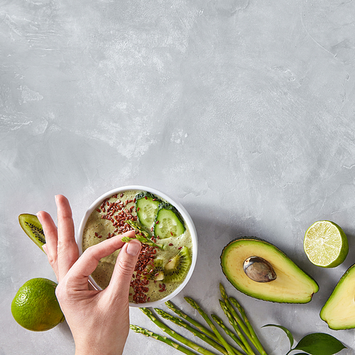 Homemade fresh smoothies from avocado, asparagus, spinach with pieces of kiwi, cucumber and flax seeds on a gray concrete background with copy space. A woman's hand puts asparagus in a plate. Dietary food. Top view
