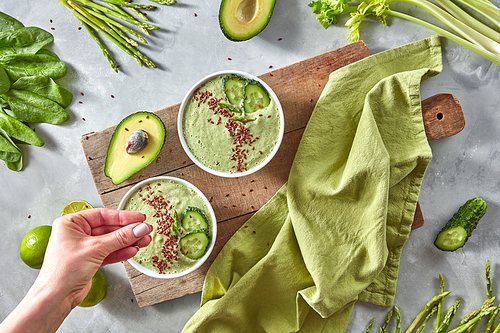 Freshly prepared homemade healthy smoothies from cucumber, avocado, spinach, lime, asparagus on a wooden board on a gray kitchen table. A woman's hand puts flax seeds in smoothies. Top view