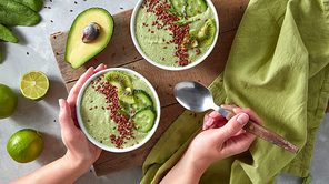 The woman's hands hold a spoon and a plate with healthy smoothies from cucumber, asparagus, avocado with pieces of kiwi and flax on a gray kitchen table. Top view