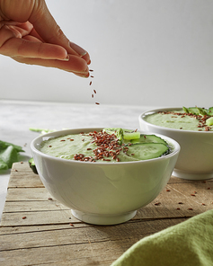 Vegetable freshly prepared smoothies with avocado, kiwi, cucumber and granola on a wooden kitchen board. A woman's hand adds flax seeds to a plate with smoothies. Healthy Eating