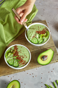 A woman's hand with a spoon, takes a vegetable smoothie. Healthy breakfast of avocado, cucumber, kiwi and flax seeds in a plate on a wooden board on a gray concrete background