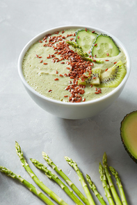 Vegetarian healthy smoothies from green organic vegetables with flax seeds and sprigs of asparagus and avocado part in a white bowl on a grey background, table.