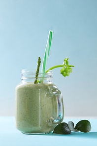 Vegetarian healthy smoothies from green vegetables with green leaves and plastic straw in a glass bowl on duotone blue paper background.
