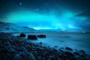 Amazing aurora borealis. Northern lights in Teriberka, Russia. Starry sky with polar lights and clouds. Night winter landscape with aurora, sea with stones in blurred water, snowy mountains. Travel