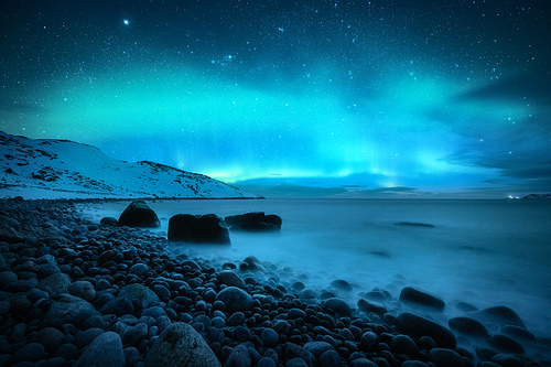 Amazing aurora borealis. Northern lights in Teriberka, Russia. Starry sky with polar lights and clouds. Night winter landscape with aurora, sea with stones in blurred water, snowy mountains. Travel