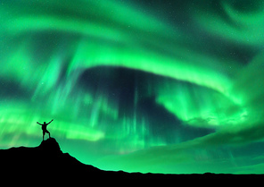 Aurora borealis and silhouette of a man with raised up arms on the mountain peak. Lofoten islands, Norway. Aurora and happy man. Sky with stars and polar lights. Night landscape with aurora and people