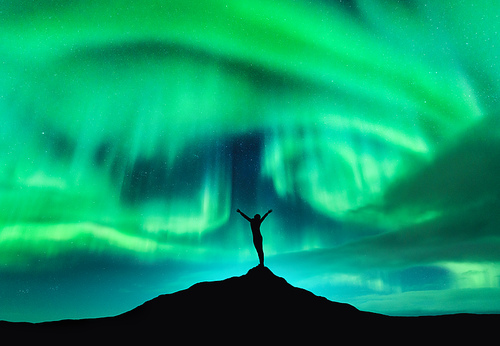 Aurora borealis and silhouette of a woman with raised up arms on the mountain peak. Lofoten islands, Norway. Aurora and happy girl. Starry sky and polar lights. Night landscape with aurora and people