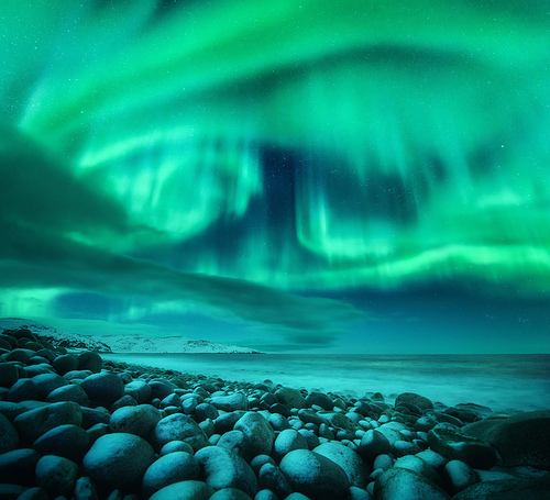 Aurora borealis over ocean. Northern lights in Teriberka, Russia. Starry sky with polar lights and clouds. Night winter landscape with beautiful aurora, sea with frozen stones in blurred water. Travel