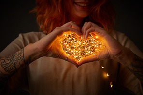 A smiling girl with a tattoo is holding in her hands a bright garland in the shape of a heart around a dark background. Happy Valentine's Day