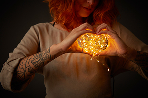 A woman is holding a yellow garland in the shape of a heart around a dark background. Layout for valentine's day