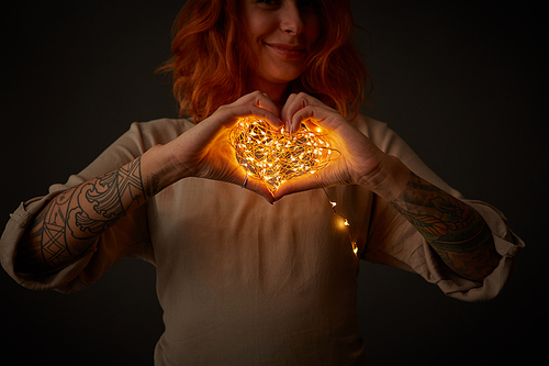 A cute girl with a tattoo is holding in her hands a bright garland in the shape of a heart around a dark background. St. Valentine's Day