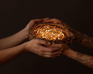 A nest of branches with bright garlands are held by male and female hands with a tattoo on a black background. Celebration