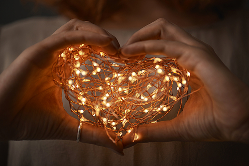 Female hands in the form of a heart hold a Christmas string of lights on a dark background