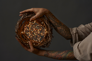 Female hands with tattoos hold a festive natural wreath of branches with a Christmas garland on a dark background