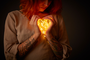 A cute girl with a tattoo is holding yellow garland lights in the shape of a heart in the dark. St. Valentine's Day