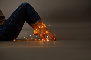 A young girl in jeans sits on the floor her legs are decorated with Christmas garlands, on a dark background with copy space for text.