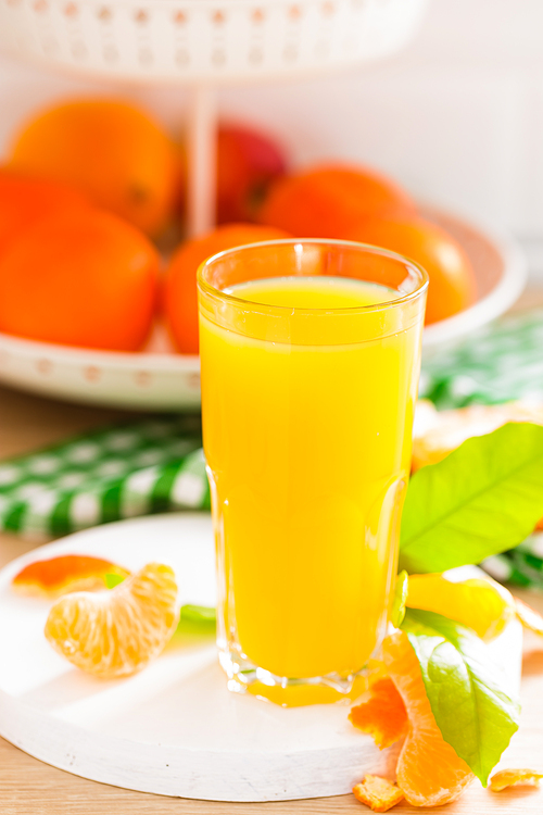 Tangerine orange juice in glass and fresh fruits with leaves on white wooden kitchen background closeup. Healthy and tasty refreshing summer beverage