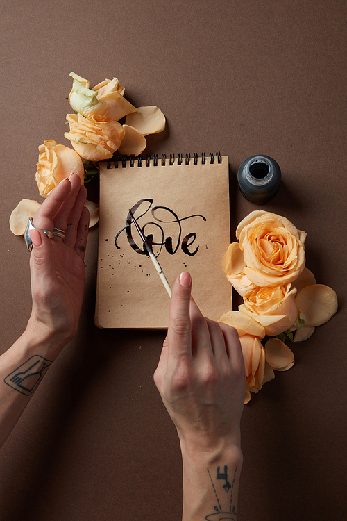 Valentine's Day concept. Notebook represented on brown background with orange flowers. Closeup of female's hand writing word love in diary or notebook in Valentine's Day.