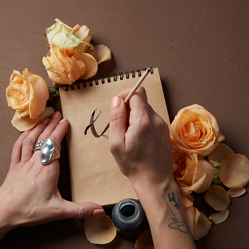 Female hands holding pen and blank paper sheets with flowers on a brown table, copyspace, flat lay