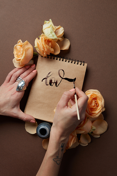 Woman writing on blank paper sheets the word love on a brown background with a bunch of flowers. Top view.