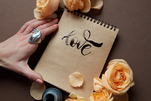 Closeup of diary or notebook with word love over brown background. Female hand not far from diary covered by orange roses.