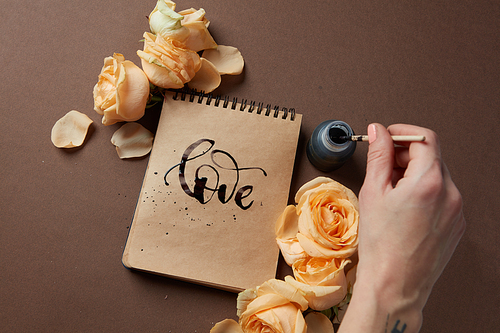 Female's hand using ink for writing word love in diary or notebook in Valentine's Day. Notebook with brown pages represented with orange flowers.