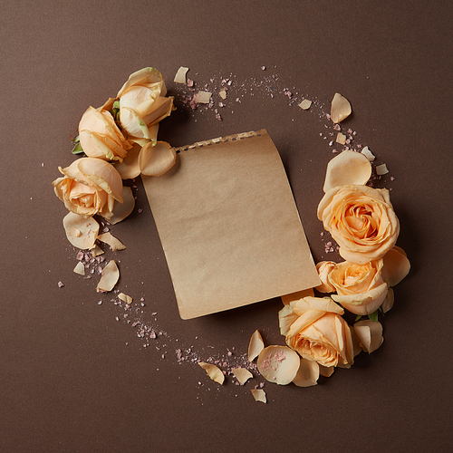Round frame of roses on a brown background with a piece of paper for your text, flat lay