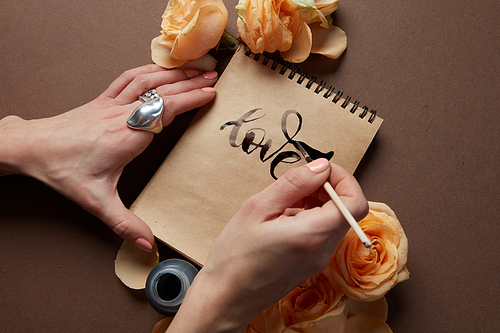 Closeup of female's hand writing word love in diary or notebook in Valentine's Day. Notebook represented on brown background with orange flowers.