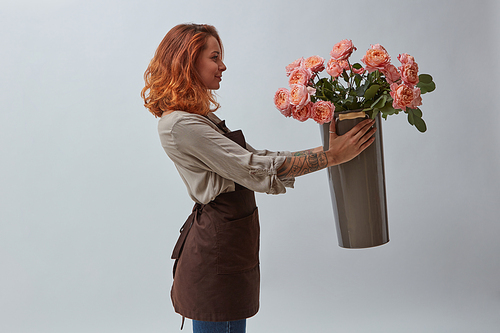 A smiling woman with a tattoo on her arm is holding a vase with pink media roses on a gray background. Happy Mother's Day
