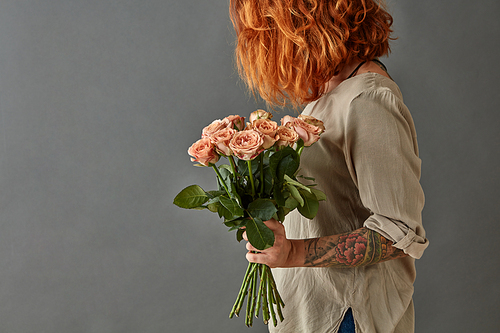 Young red-haired girl with a tattoo holds a bouquet of pink roses on a gray background
