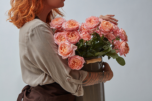 A bouquet of fresh pink roses in a vase is held in hands by a red-haired girl florist in a brown apron around a gray background with copy space.